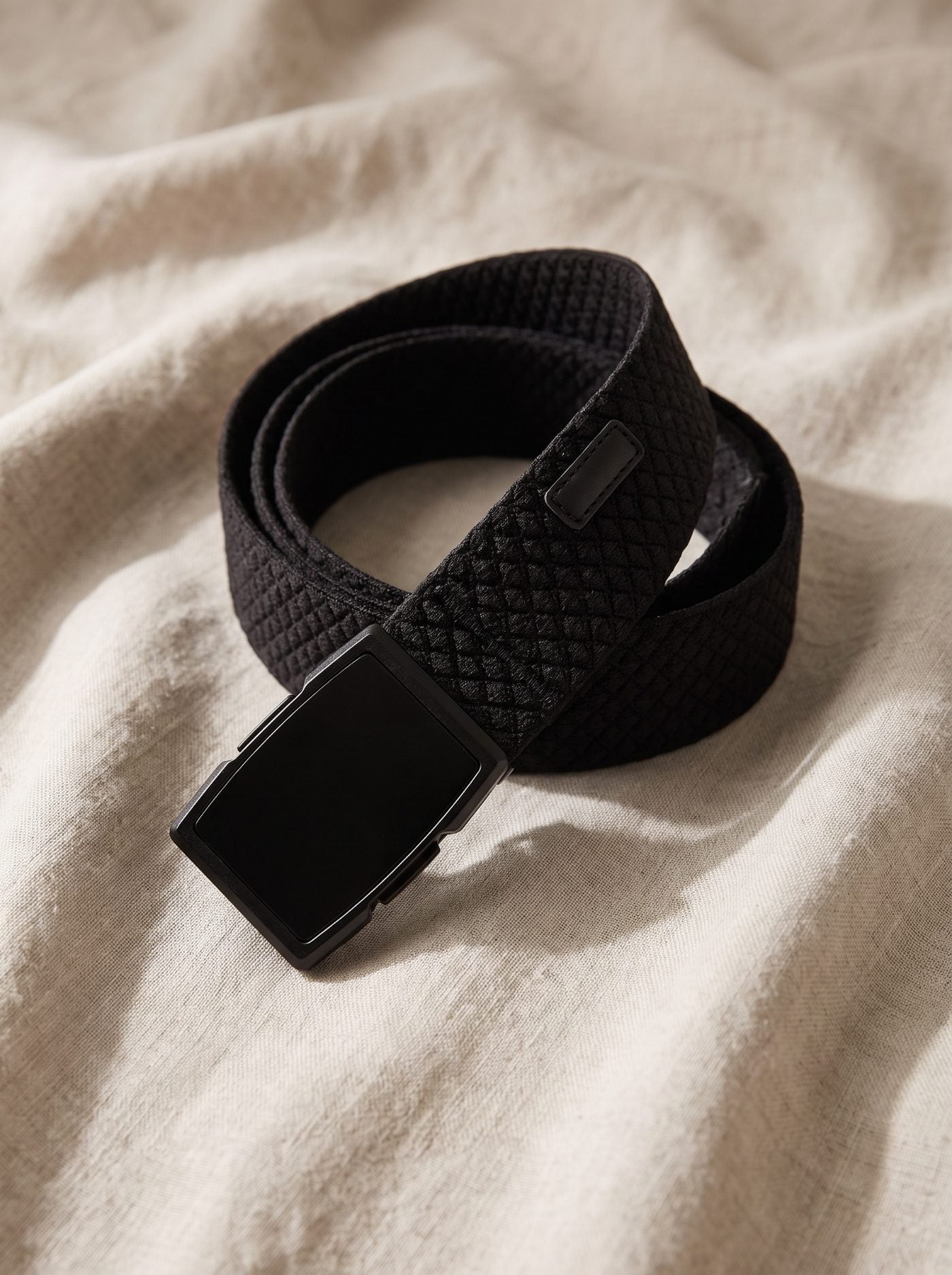 The Flexy belt coiled on natural linen, showing the diamond weave of the webbing and the matte black buckle