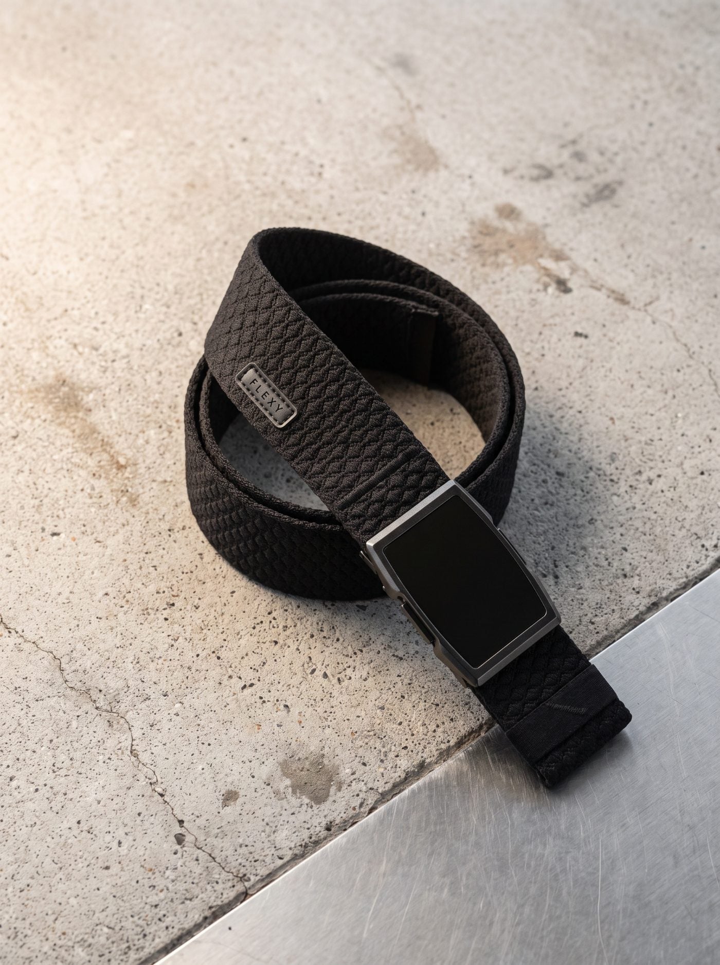 The Flexy belt resting on a concrete ledge, buckle open, with the woven Flexy tab visible on the strap